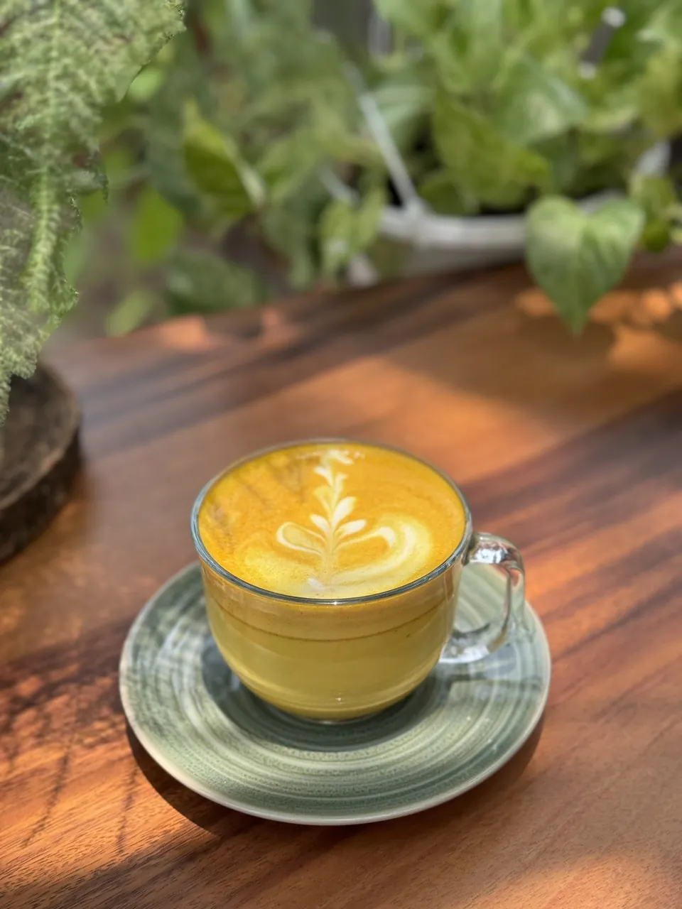 Un golden milk latte con arte en la espuma servido en un vaso amarillo, con el jardín desenfocado detrás