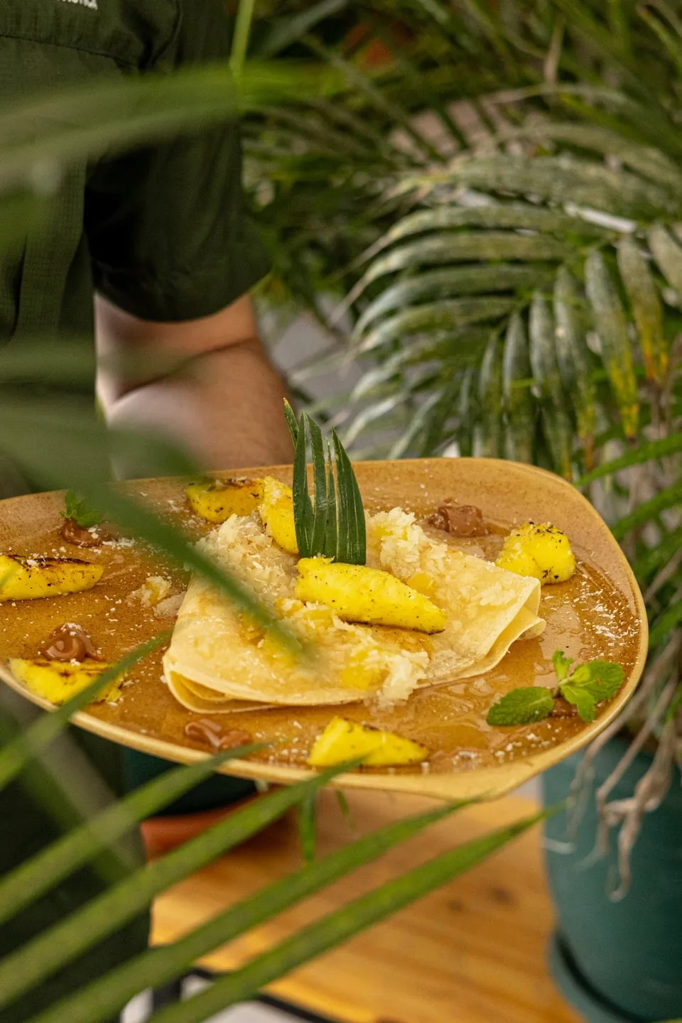 Un mesero sostiene una crepa de piña y coco entre hojas de palma del jardín