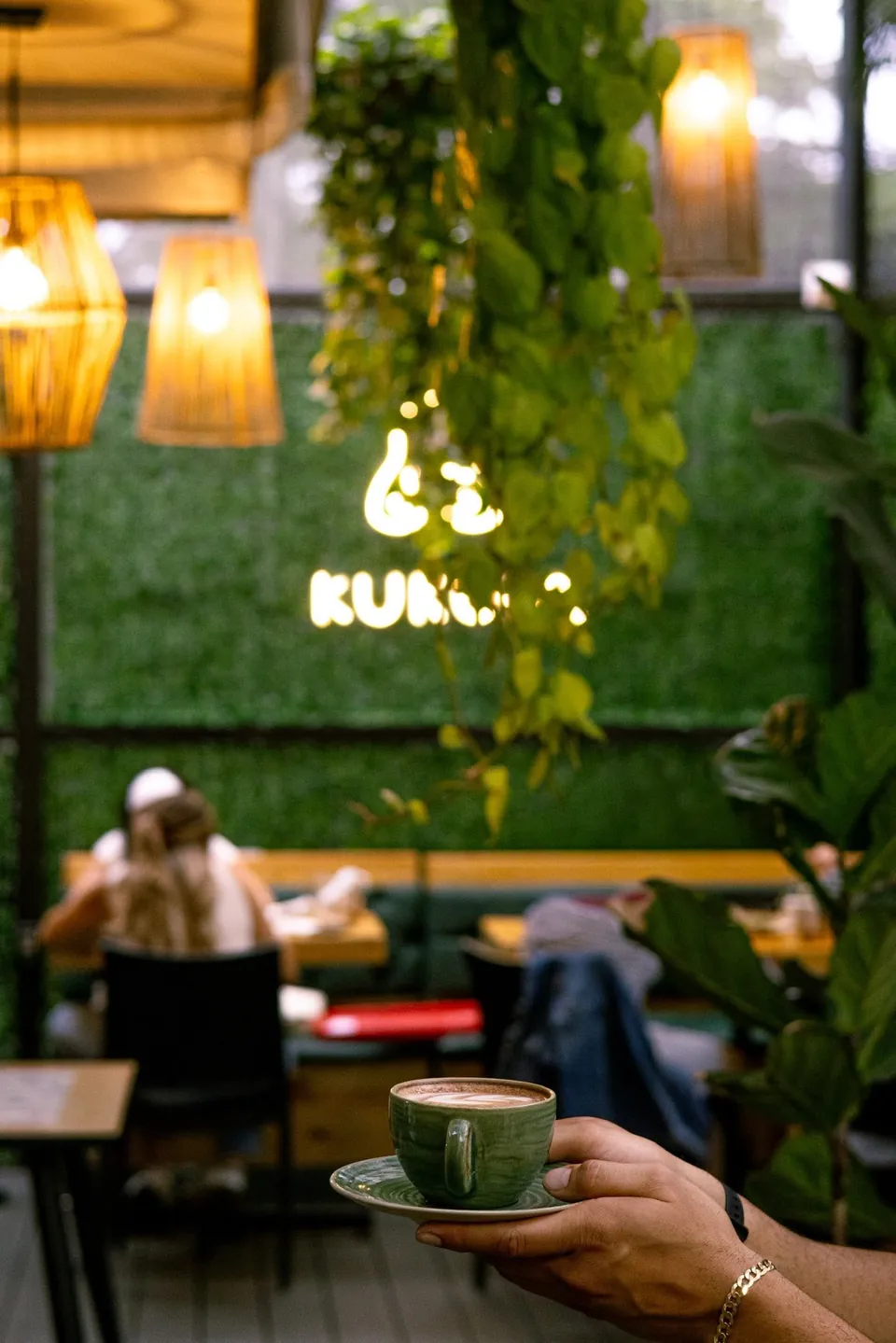 Una mano sostiene un latte verde de especialidad frente al rótulo de neón del perezoso Kukula, entre plantas del jardín