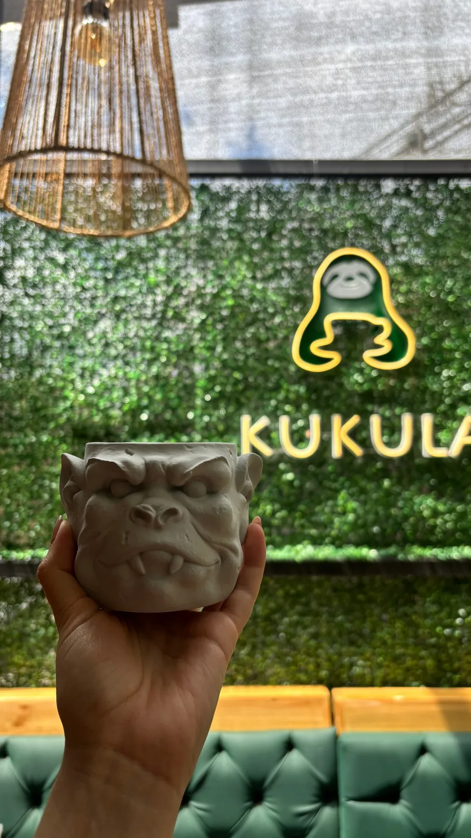 Una mano sostiene una taza de gárgola sin pintar frente al neón encendido del perezoso Kukula
