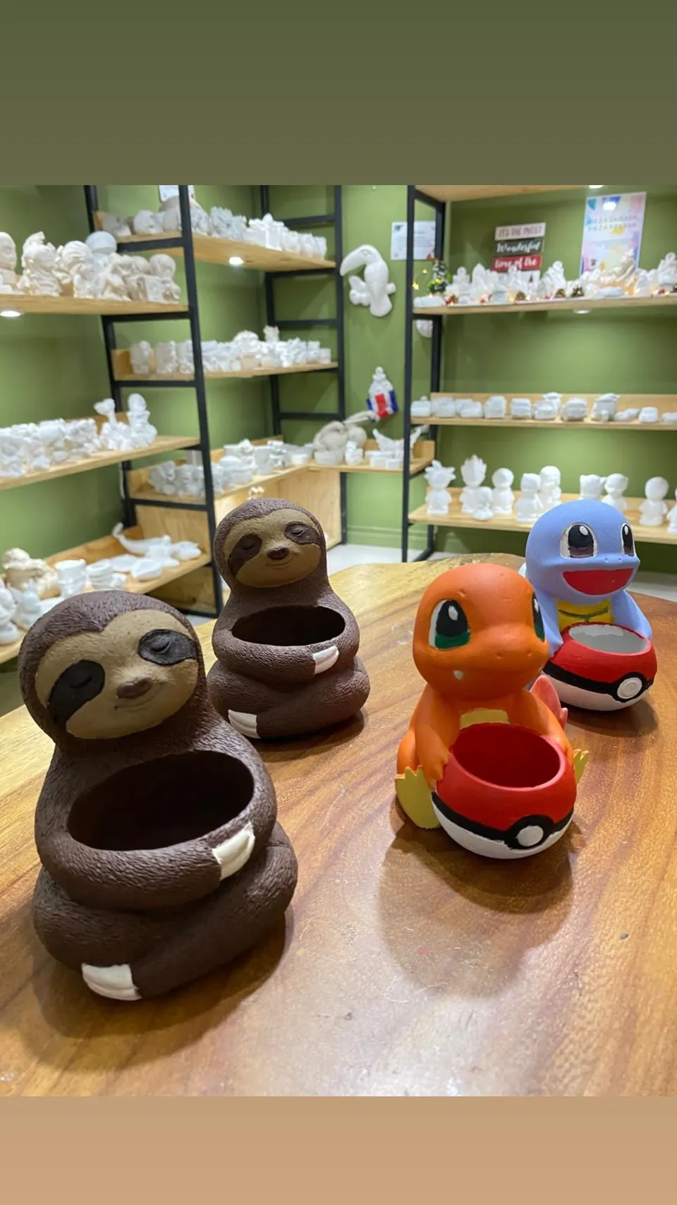 Maceteros con forma de perezoso pintados a mano junto a figuras de Pokémon, frente a estantes de cerámica sin pintar