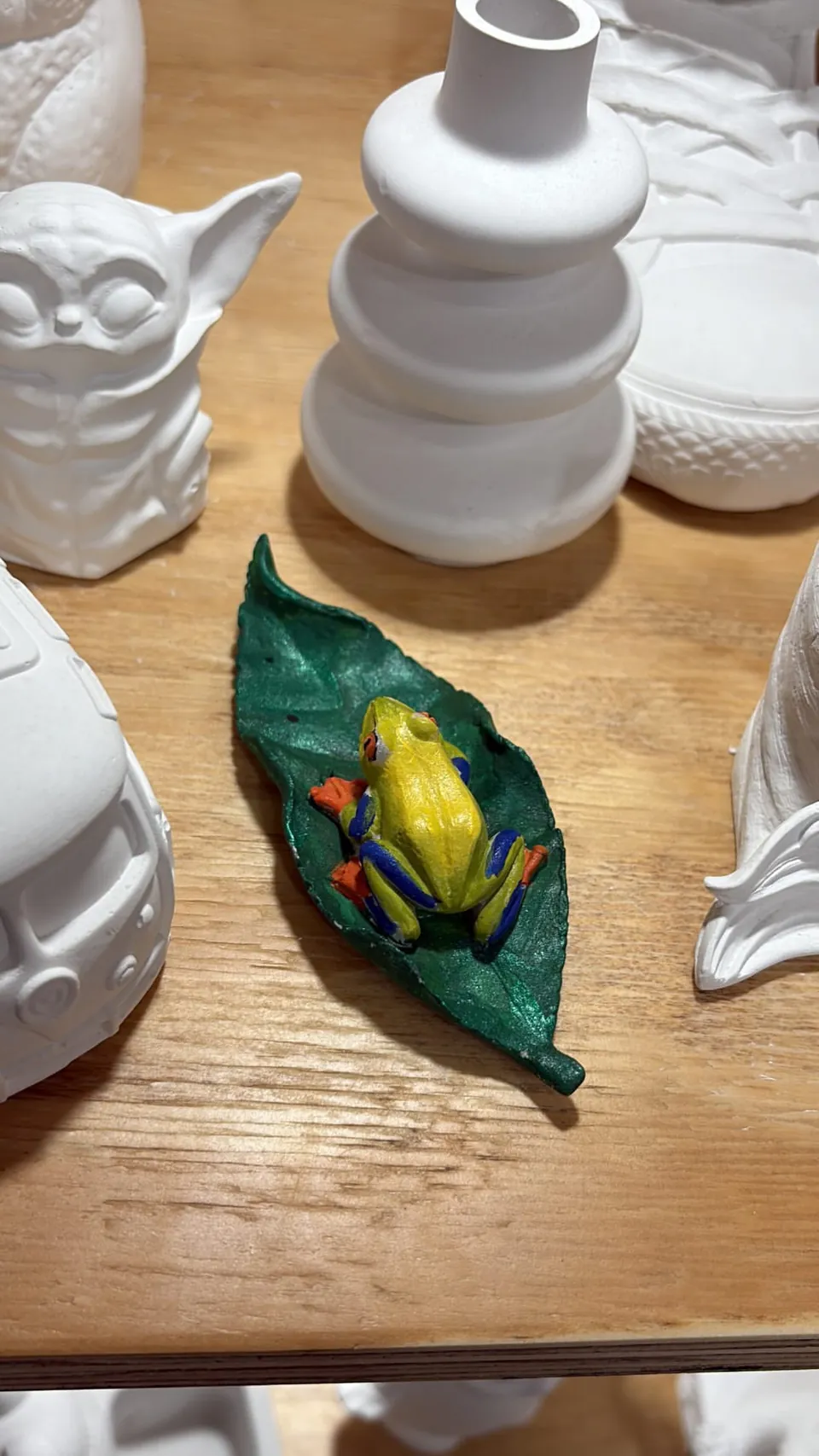 Una rana de ojos rojos pintada sobre un plato con forma de hoja, entre cerámica blanca